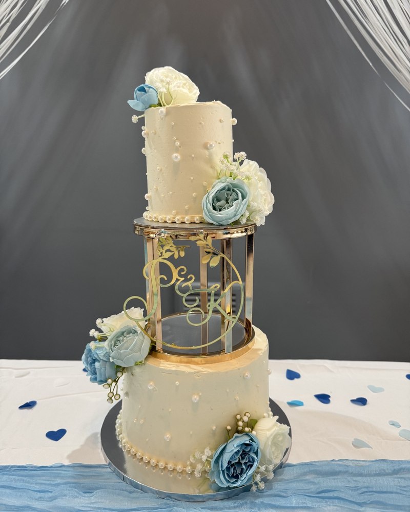 Two-tier wedding cake with gold birdcage stand and dusty blue florals, P&K monogram